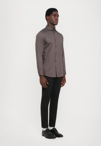 Herre langermet skjorte i mørk brun, med et teksturert geometrisk mønster, knappestolpe krage og slim fit. Kombinert med svarte bukser og sko.