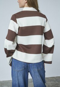 Langærmet, skjorte med krave, der har iøjnefaldende vandrette brune og hvide striber. Stoffet ser ud til at være tekstureret; parret med løse denimbukser.