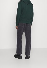 Sweat à capuche vert et jean large gris foncé, porté avec des bottes noires épaisses. Style décontracté avec des coutures visibles sur le pantalon.