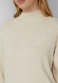 Beżowy dziergany sweter z wysokim kołnierzem, z równomiernie rozmieszczonymi dekoracyjnymi perłami na przodzie. Gładka tekstura i luźny krój.
