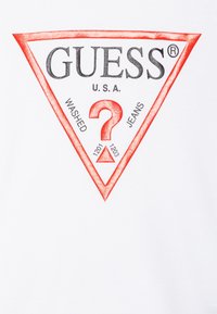 Tessuto bianco con un logo triangolare rosso con la scritta "GUESS" e "U.S.A.", un punto interrogativo centrale, e il testo aggiuntivo "WASHED JEANS" e numeri.