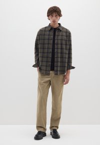 Camicia a quadri in tonalità di verde e marrone, indossata sopra un collo tondo nero, abbinata a pantaloni beige e scarpe nere con texture.