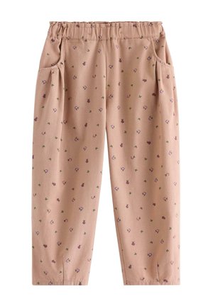Junges rothaariges Mädchen lächelt, trägt eine beige gesteppte Weste, ein rosa Langarmshirt, gemusterte beigefarbene Hose und braune gewebte Schuhe, steht mit den Händen in den Taschen.