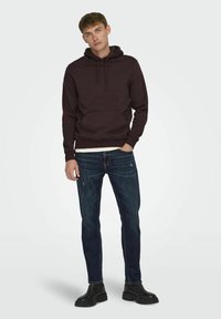 Sudadera marrón con capucha y bolsillo frontal, combinada con jeans oscuros desgastados. La modelo lleva botas negras hasta el tobillo. Diseño simple, material de algodón.