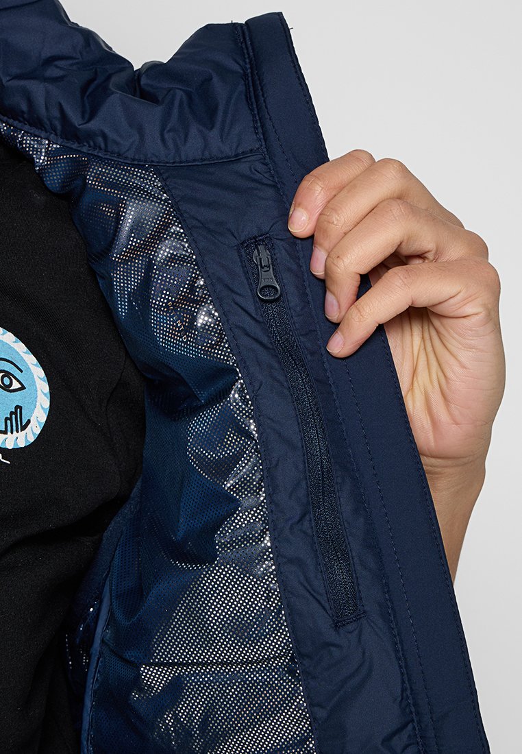 Veste bleu marine avec une finition lisse, dotée d'une poche zippée et d'une doublure intérieure brillante et à motifs. Main tenant l'ouverture de la veste.