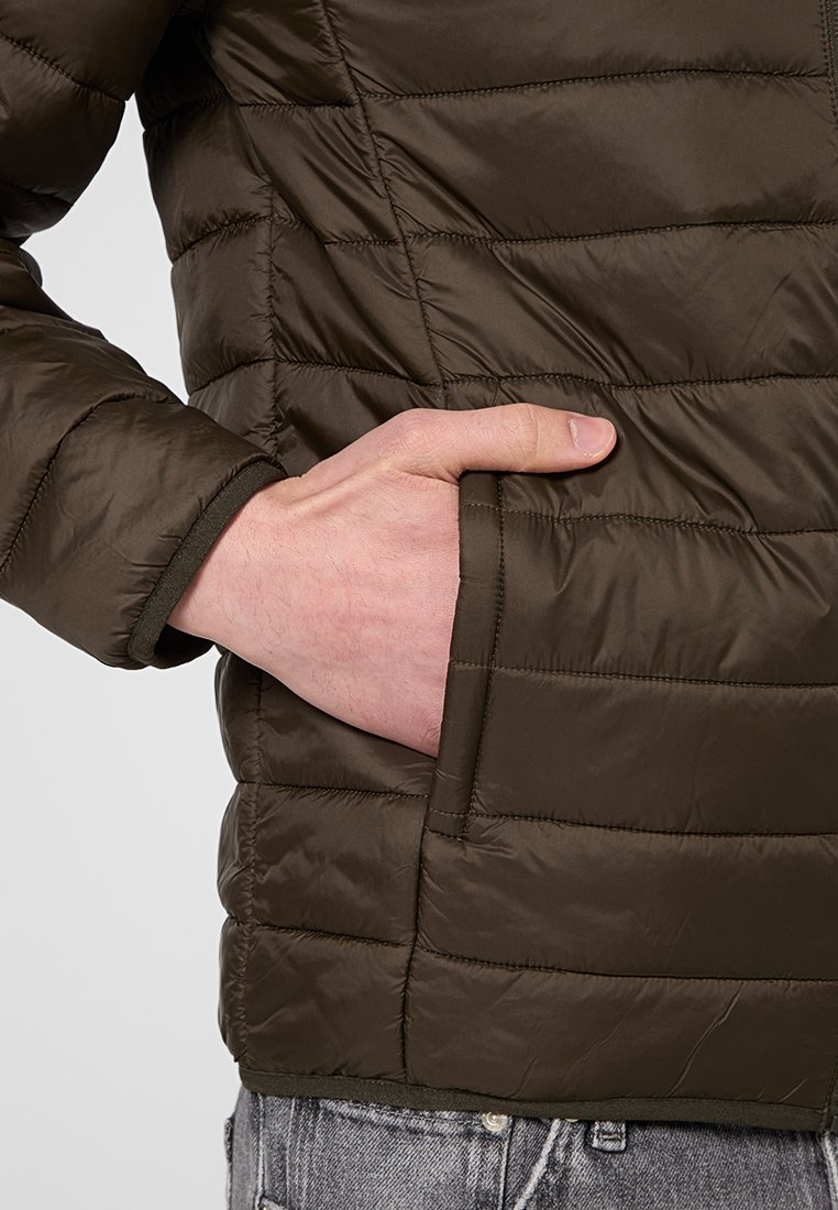 Veste matelassée vert olive foncé avec des sections matelassées, une poche zippée et des cuffs côtelés pour plus de confort et d'isolation.