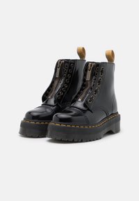 Stivaletti in pelle verniciata nera con doppie cerniere, dettagli in metallo dorato, suola in gomma nera con texture e cuciture gialle.