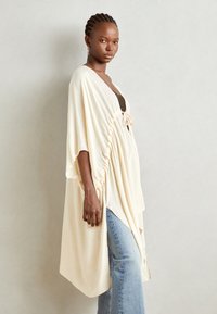 Lys kremfarget kimono-lignende omslag med knytelukking, myk tekstur, avslappet passform og side-ruffle. Brukt over en mørk topp og denimjeans.