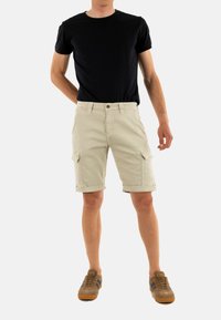 Pantaloni cargo beige con tasche frontali e laterali, orlo risvoltato e chiusura con bottone, abbinati a una maglietta nera e sneakers marroni.