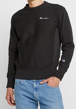 Mann trägt schwarzen Champion-Sweatshirt mit Logo auf Brust und Ärmel, kombiniert mit hellblauen Jeans, Hand in der Tasche.