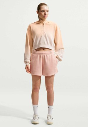 Junge Frau steht und trägt einen hellpfirsichfarbenen kurzen Hoodie, pinke Shorts, weiße Socken und Turnschuhe vor hellem, einfarbigem Hintergrund.