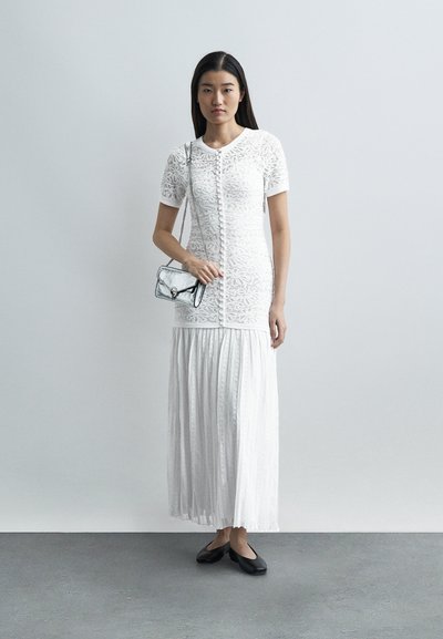 Mladá žena stojí opřená o jednoduchou zeď, má na sobě bílý krajkový vršek a plisovanou sukni, drží malou stříbrnou kabelku a má černé baleríny.