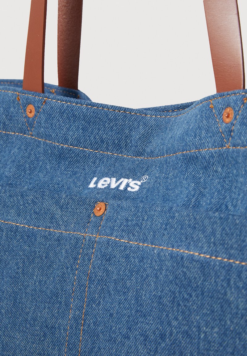 Denim draagtas met bruine faux leren handvatten, zichtbare stiknaden en Levis merklogo in het wit. Bevat koperen hardwareaccenten.