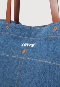 Denim torba za nakupovanje z rjavimi umetnimi usnjenimi ročaji, vidno šivanjem in Levis blagovno znamko v belem. Opremljena je s koprenastimi kovinskimi dodatki.