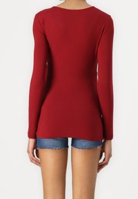 Czerwona bluzka z długim rękawem, dopasowany krój, okrągły dekolt, rozciągliwy materiał, w zestawie z dżinsowymi szortami. Bez zauważalnych wzorów ani detali metalowych.