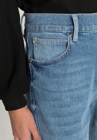 Jeans de denim azul claro com uma cintura alta, apresentando um único bolso traseiro, costura em contraste e um tecido liso e texturizado.