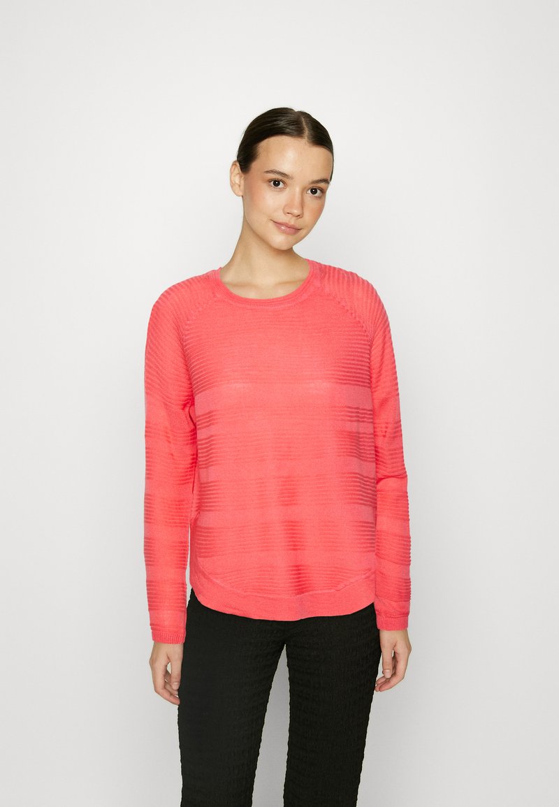 Pull long à manches longues de couleur corail avec un motif texturé en côtes, encolure ronde et coupe décontractée, associé à un pantalon noir texturé.