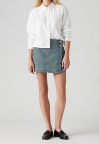 Camisa blanca de manga larga con botones combinada con una falda de mezclilla color teal con diseño asimétrico y cierre de botones en el costado. Mocasines negros completan el look.