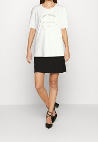 Camiseta de algodón blanca con cuello redondo y mangas cortas, con un sutil estampado dorado. Se lleva con una mini falda negra y tacones negros.