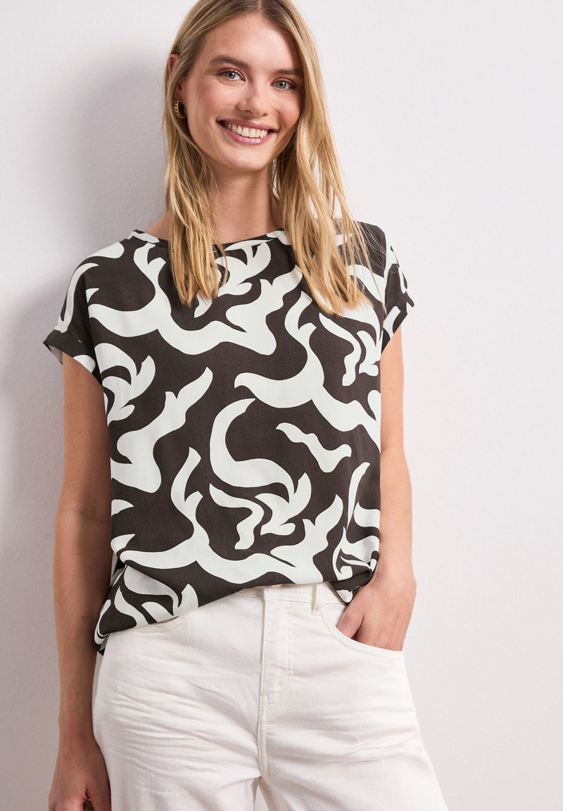 Mujer sonriente con cabello largo y rubio que lleva una blusa con estampado abstracto en blanco y negro y pantalones blancos, con la mano en el bolsillo, frente a una pared lisa.