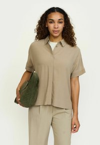 Kortærmet, knap-op skjorte i lysebrun stof, parret med beige bukser, og holder en olivengrøn ruskinds taske. Glat tekstur, afslappet pasform.