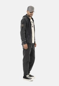 Hoodie gris avec un patch logo, associé à un t-shirt crème, un jean noir et des baskets noires avec des semelles blanches. Homme portant une casquette grise.