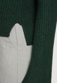 Pull vert en tricot avec un motif texturé, présentant une coupe courte et un large col, porté avec un tissu gris clair en dessous.