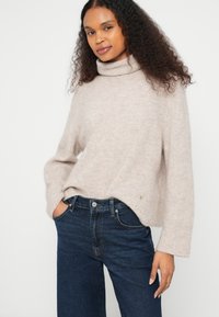 Mulher com cabelo encaracolado vestindo um suéter de gola alta bege e jeans azul escuro, em pé com uma mão no bolso contra um fundo liso.