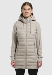 Beige hybridjakke med en fyldig quiltet krop og bløde stofærmer, med hætte, fuld lynlås og to sidelommer.