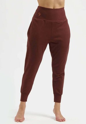 Bruine joggerbroek met hoge taille, elastische boorden en zijzakken, gedragen op blote voeten tegen een lichte achtergrond.