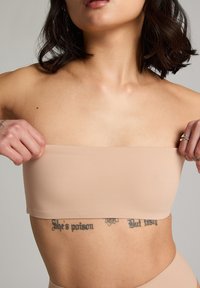 Nude strapless top gemaakt van een gladde stof. Heeft een eenvoudig ontwerp en naadloze randen. Model past de top met één hand aan.