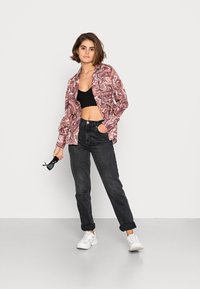 Camisa com padrão abstrato multicolorido, top cropped preto, jeans escuros de cintura alta e ténis brancos. Óculos de sol segurados em uma mão.