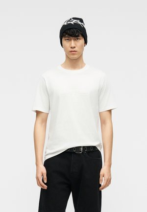 Νεαρός άνδρας που φοράει λευκό κοντομάνικο μπλουζάκι, μαύρο παντελόνι με μαύρη ζώνη και μαύρο σκουφί με λευκό αστέρι.