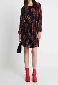Abito nero plissettato con maniche lunghe, decorato con motivi floreali rossi e rosa, abbinato a stivali con tacco rossi e una borsa nera.