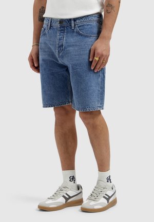 Man draagt blauwe denim shorts, witte sokken met zwart logo, witte en grijze sneakers en ringen aan de rechterhand, staand op een effen grijze achtergrond.