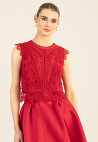 Rotes Spitzenoberteil mit gewellten Ärmeln und komplexen Blumenmustern, kombiniert mit einem glatten roten Rock, der an der Taille Falten hat.