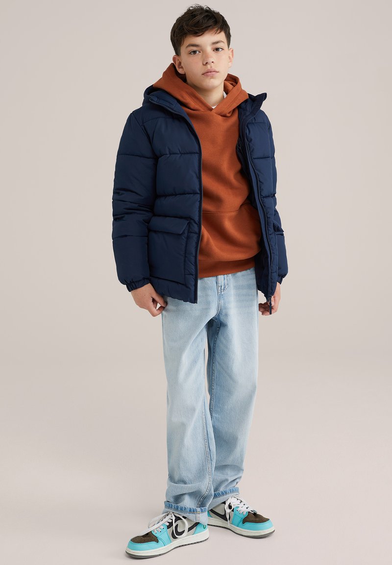 Giubbotto imbottito blu marino con cappuccio, felpa marrone sotto, jeans azzurri chiari e sneaker multicolori con accenti neri e turchesi.