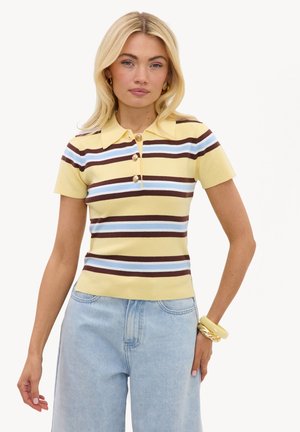 Mujer rubia que lleva una camisa polo de manga corta con botones a rayas amarilla, azul y marrón y vaqueros de cintura alta de color azul claro, posando con la mano en la cadera.
