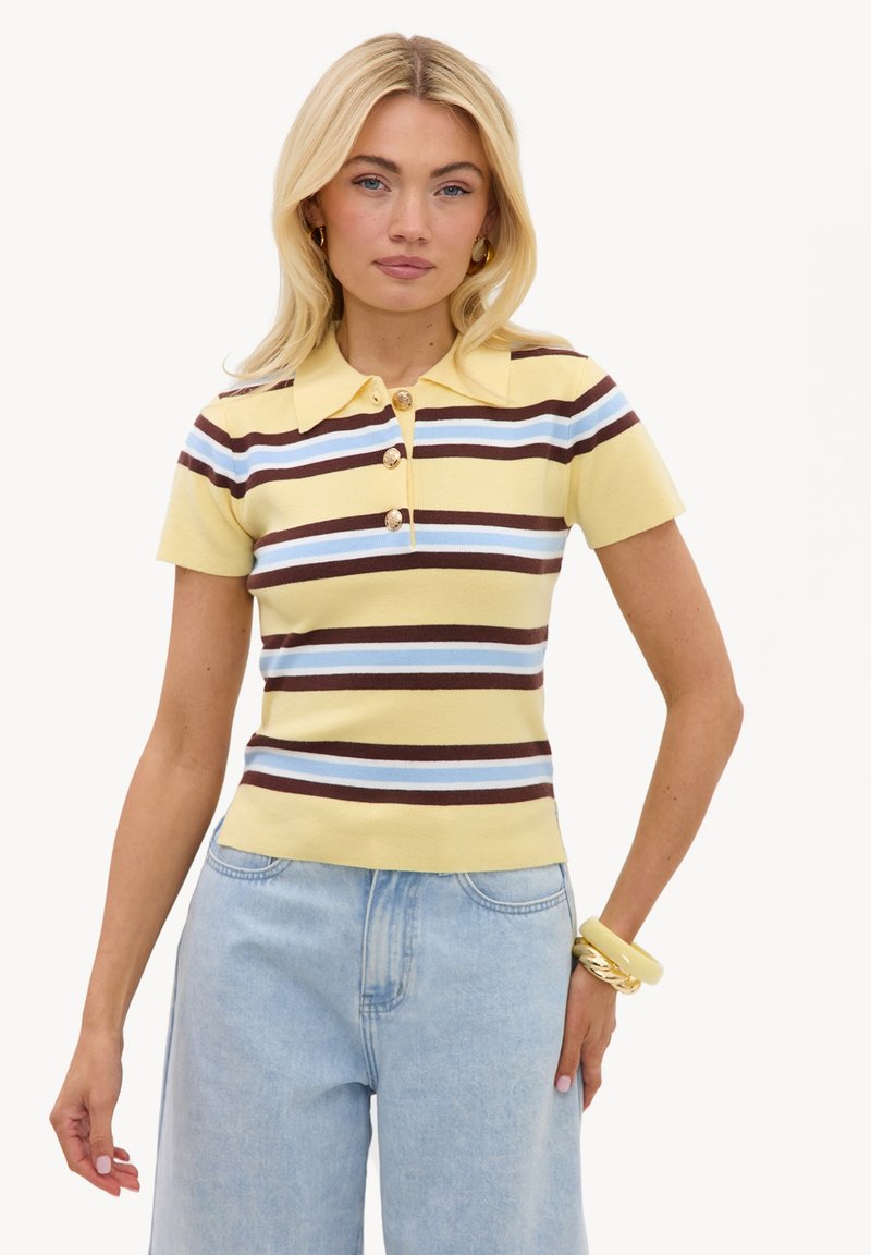 Femme blonde portant un polo à manches courtes rayé jaune, bleu et marron avec des boutons et un jean taille haute bleu clair, posant la main sur la hanche.