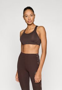 Hnedá športová podprsenka so širokými ramienkami, s hladkou textúrou a jemným logom. Kombinovaná s vysokými legínami v rovnakom prevedení.
