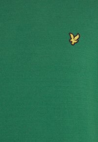 Zaļš kokvilnas T-krekls ar dzeltenu izšūtu putna logotipu uz krūtīm, ar gludu, ribotu tekstūru.