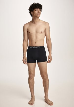 Young man with curly hair standing barefoot, wearing black Jockey boxer briefs, against a plain light background.