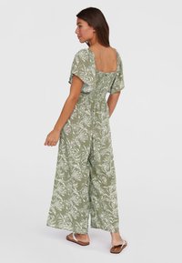 Combinaison verte avec un motif floral blanc, dos carré, manches courtes, taille élastique et jambes larges. Texture lisse ; associée à des sandales plates.