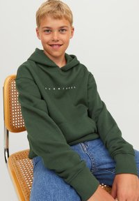 Sudadera verde con capucha y un bolsillo delantero, con el texto blanco "ACK & JONES". Llevada con jeans azules, sentado en una silla de caña.