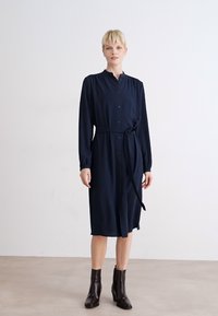 Navy blue, long-sleeve shirt dress with a button-down front, tie waist, and round collar. Smooth texture, paired with dark ankle boots.