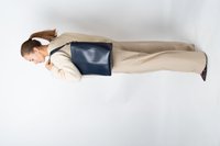 Mujer con coleta, vestida con un suéter beige y pantalones, llevando una gran bolsa de piel azul marino al hombro, de pie de lado sobre un fondo blanco.