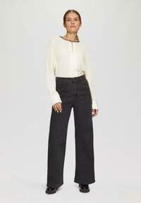 Blouse à manches longues crème avec un col décoratif, portée au-dessus d'un jean noir taille haute à jambes larges. Tissu lisse et légère brillance sur les chaussures.