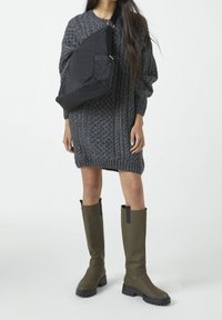 Robe pull en maille grise avec un motif texturé, associée à des bottes vert olive montantes avec des accents noirs, et un sac à épaule noir.