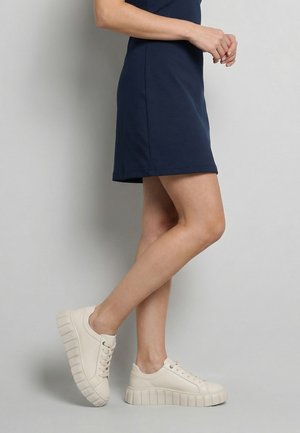 Vrouw draagt een marineblauwe jurk en beige plateau sneakers, staand met één voet lichtjes opgetild tegen een effen grijze achtergrond.