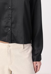 Persona che indossa una camicia nera a maniche lunghe con bottoni, infilata in pantaloni beige a vita alta con chiusura a bottone e passanti per cintura.